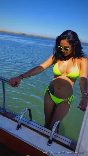 Woman in bright green bikini standing on boat railing by water, wearing sunglasses.