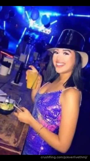 Woman in sparkly purple top and hat holding drink at bar with blue lights.