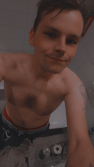Shirtless man with tattoos and necklace in bathroom, wearing swim trunks.