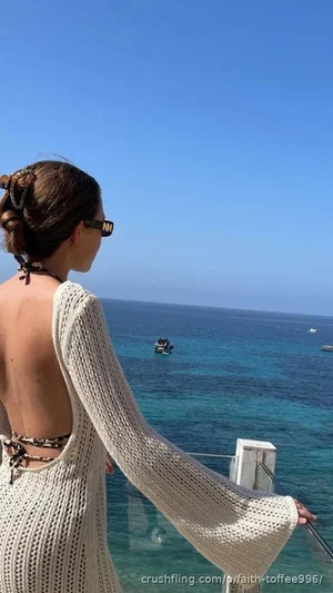 Woman in white knit dress with open back, standing by ocean, wearing sunglasses, hair in bun.