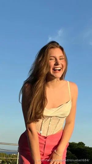 Woman with long brown hair in a beige top and pink pants, smiling outdoors under a clear blue sky.