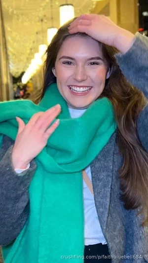 Woman with long brown hair wearing green scarf and gray coat, smiling indoors.