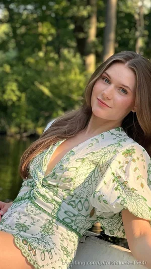 Woman in green patterned dress, long brown hair, sitting by water in forest.