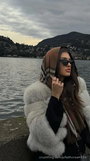 Woman in fur coat and scarf by lake, wearing sunglasses, outdoor setting.