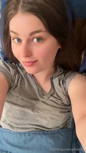 Woman with brown hair in gray shirt lying on blue bedding, indoor selfie.