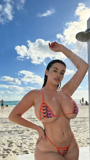 Woman in orange patterned bikini under outdoor shower on beach, posing with hand on hip.