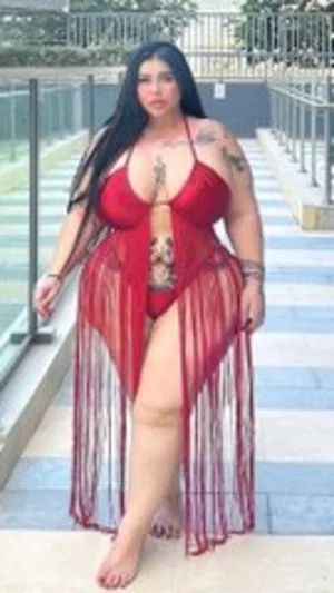 Woman in red sheer dress with tattoos, standing outdoors on a walkway.