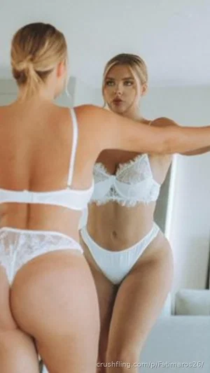 Woman in white lace lingerie poses in mirror selfie, indoor setting.