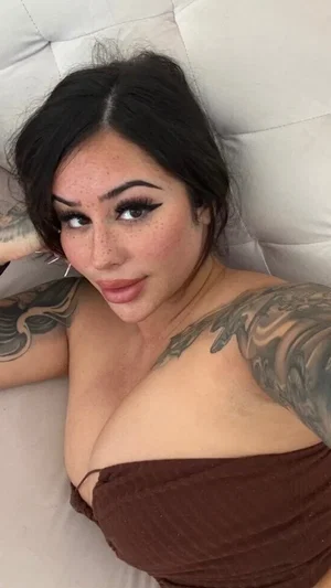 Woman with dark hair and tattoos lying on a white couch, wearing a brown top.