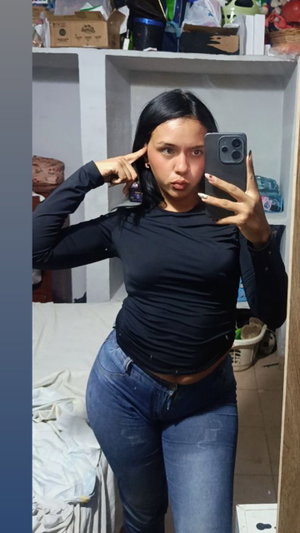 Curvy woman with black hair in black top and jeans in bedroom.