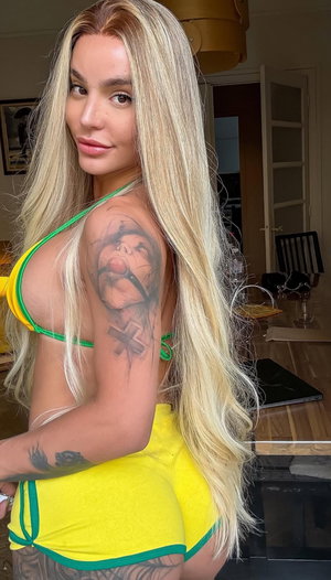 Woman with long blonde hair in yellow and green bikini, tattoos on arm and back, indoor setting.