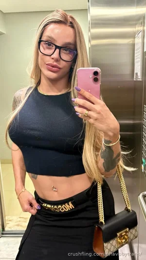 Woman with blonde hair and glasses wears black crop top and skirt, holding phone in elevator.