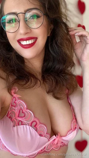 Woman with dark curly hair wearing pink lace bra and glasses, smiling in front of red heart