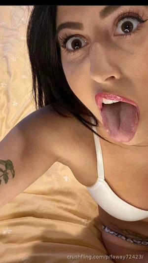 Woman with dark hair in white bra, tongue out, lying on yellow floral bedding.