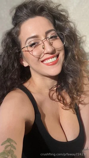 Woman with curly hair and glasses, wearing black tank top, smiling in indoor setting.