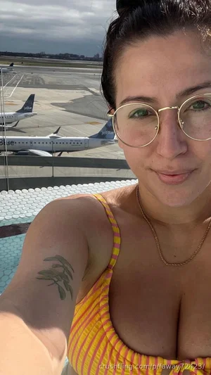 Woman in yellow striped top with glasses, tattoo on arm, at airport with planes in background.