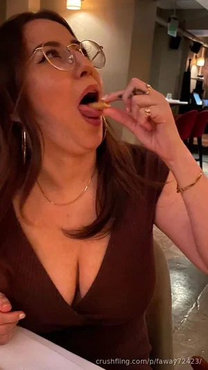 Woman with glasses eating food in a restaurant, wearing a brown top.