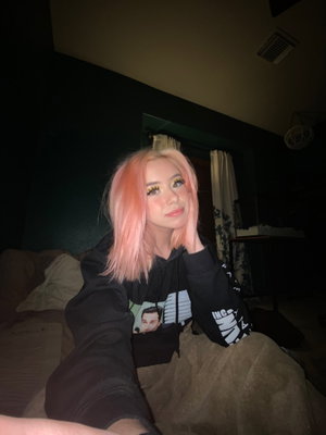 Woman with pink hair in black hoodie sitting on bed indoors.