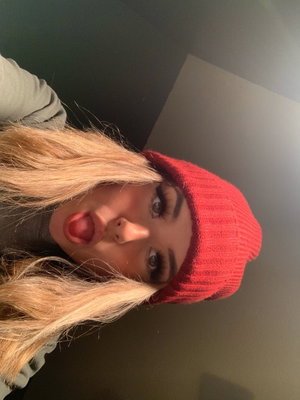 Woman with blonde hair and red beanie sticks out tongue in indoor selfie.