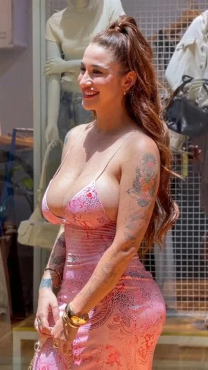 Woman in pink lace dress with tattoos, standing outdoors near storefront.