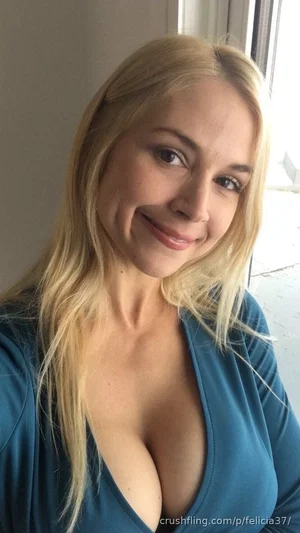Blonde woman in a blue top with a deep neckline, smiling indoors near a window.