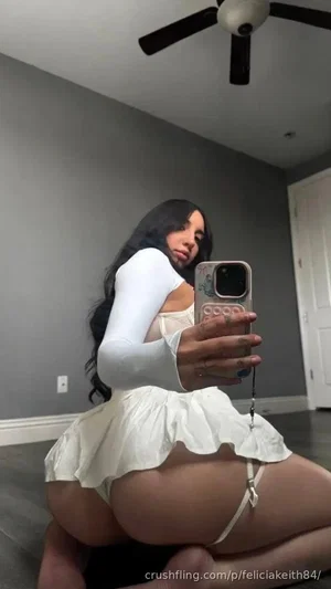 Woman with long dark hair in white long-sleeve top and skirt, kneeling indoors, holding phone for