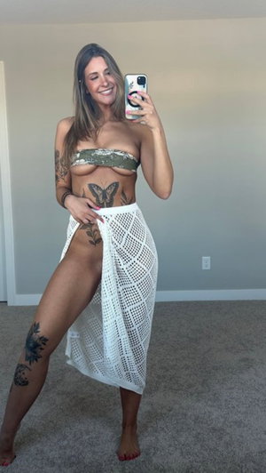 Woman with tattoos taking mirror selfie in bedroom wearing skirt.