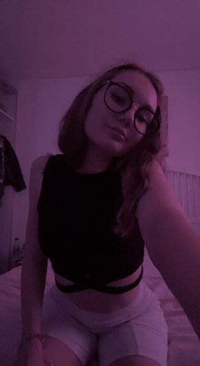 Woman with glasses in black crop top and white shorts sitting on bed in dim purple light.