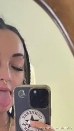 Woman with braided hair and nose ring taking mirror selfie, tongue out, holding phone.
