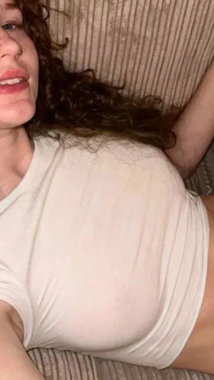 Woman with curly hair lying on couch, wearing tight white top, indoor setting.