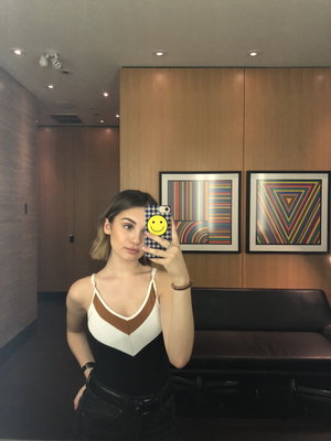 Woman in black and white top, holding phone with smiley cover, standing indoors near artwork.