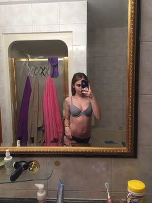 Woman in gray bra and black underwear takes mirror selfie in bathroom.