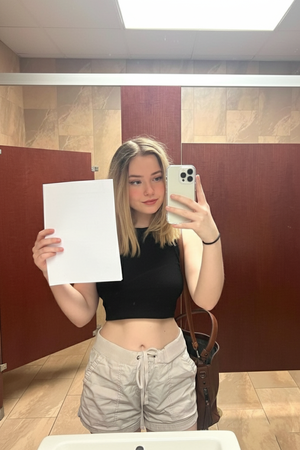 Woman with blonde hair in black crop top and beige shorts taking mirror selfie in bathroom.