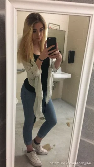 Woman with blonde hair in jeans and a black top takes mirror selfie in bathroom.