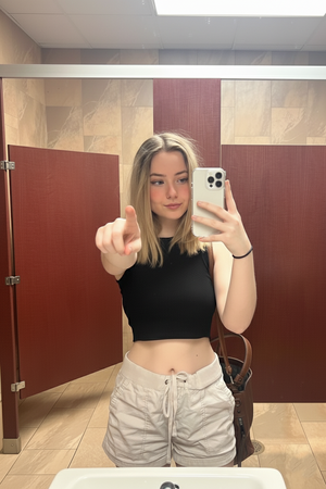 Woman with blonde hair in black crop top and light shorts taking mirror selfie in bathroom.