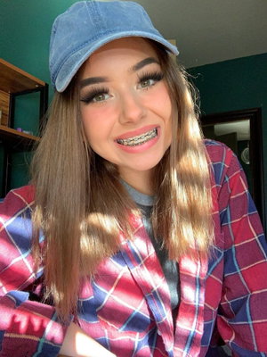 Woman with long hair, blue cap, plaid shirt, smiling with braces, indoors.