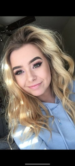 Blonde woman with wavy hair wearing a light blue top, smiling in a close-up selfie.