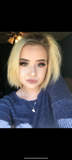 Blonde woman in blue sweater taking selfie indoors with makeup and necklace.