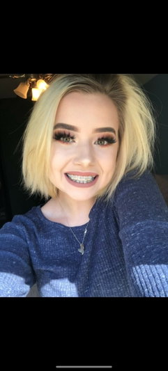 Blonde woman in blue sweater smiling, indoor selfie with warm lighting.