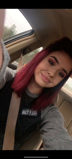 Woman with pink hair in a car, wearing a gray and black shirt, smiling at camera.