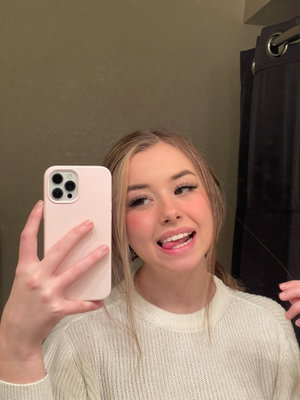 Woman with blonde hair in a white sweater taking mirror selfie in bathroom.