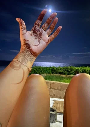Woman with henna on hand and legs, sitting outdoors at night near ocean.