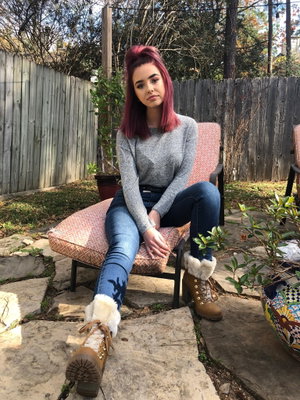 Woman with purple hair sits on patio chair, wearing gray sweater, jeans, and boots.