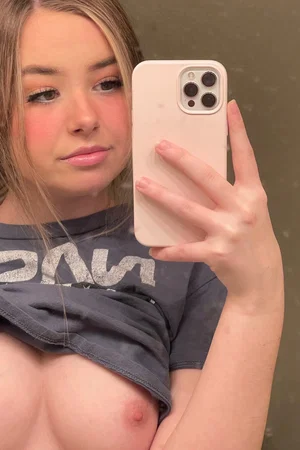 Woman with blonde hair in a mirror selfie, shirt pulled down to expose breasts, indoor setting.