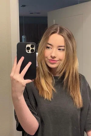 Woman with long brown hair in a dark gray t-shirt taking a mirror selfie indoors.