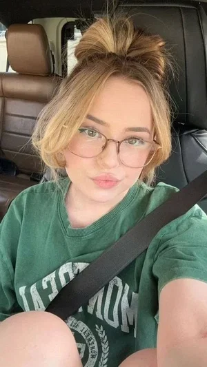 Woman with blonde hair in bun, wearing glasses and green t-shirt, sitting in car.