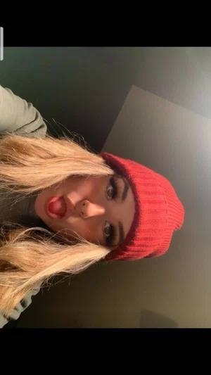 Woman with blonde hair and red beanie posing indoors, tongue out, wearing gray top.