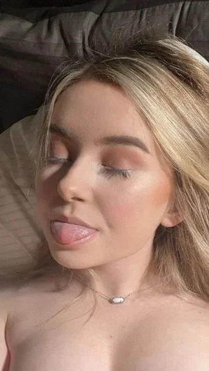 Blonde woman with makeup lying on bed, topless, sticking out tongue.