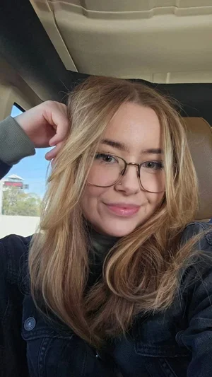 Woman with blonde hair and glasses, wearing a dark jacket, sitting in a car.