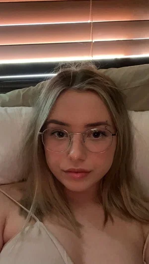Woman with blonde hair and glasses lying in bed, wearing a light-colored top.